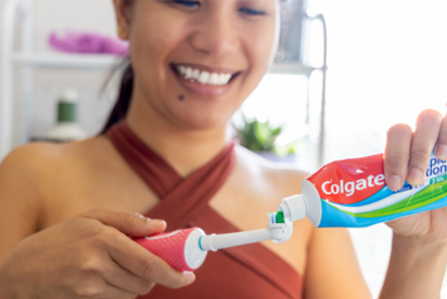 Woman smiling while adding Colgate toothpaste to a toothbrush in a bathroom.
